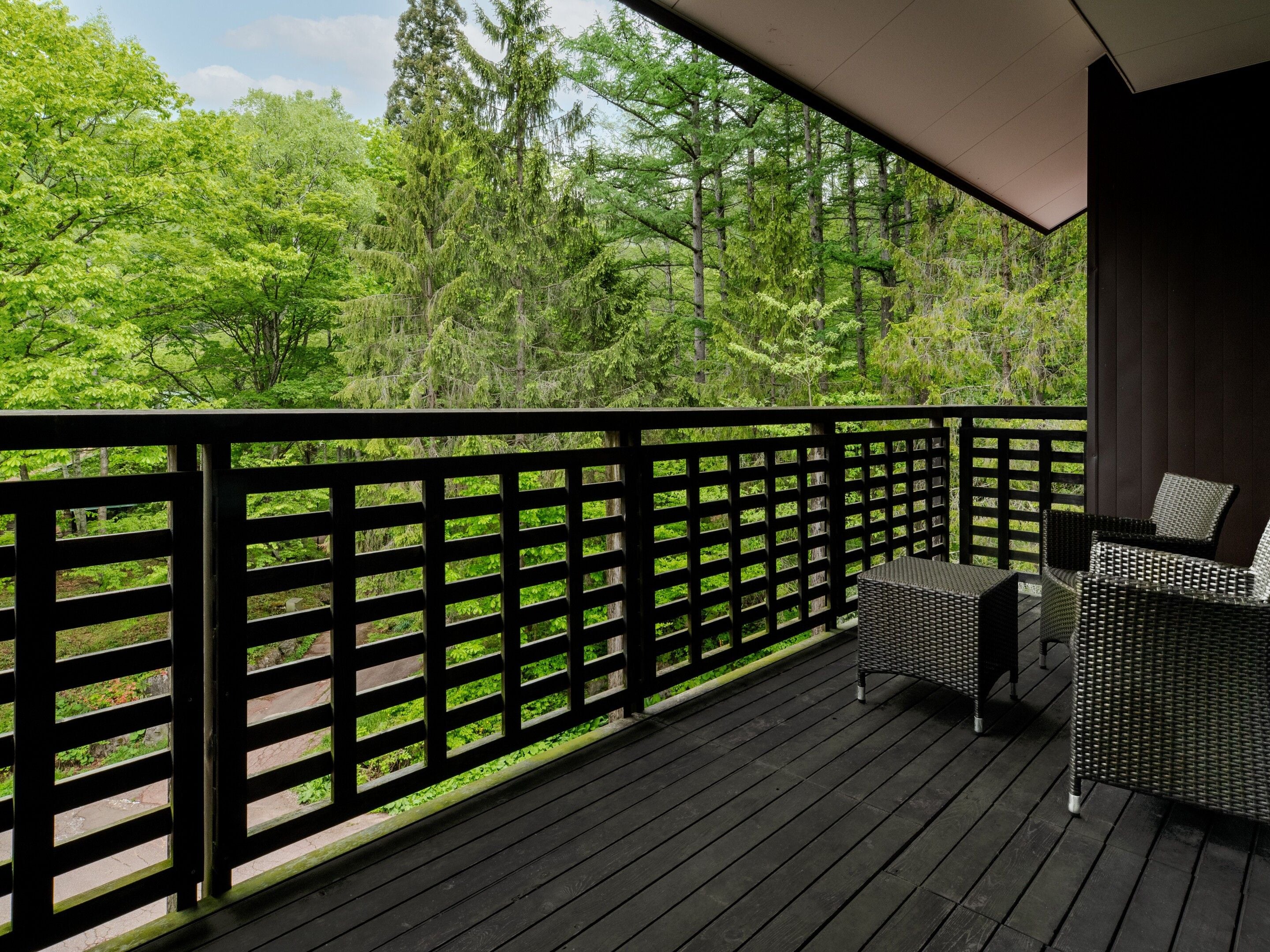 A wooden deck with a black railing, a wicker sofa, and a view of a forest.