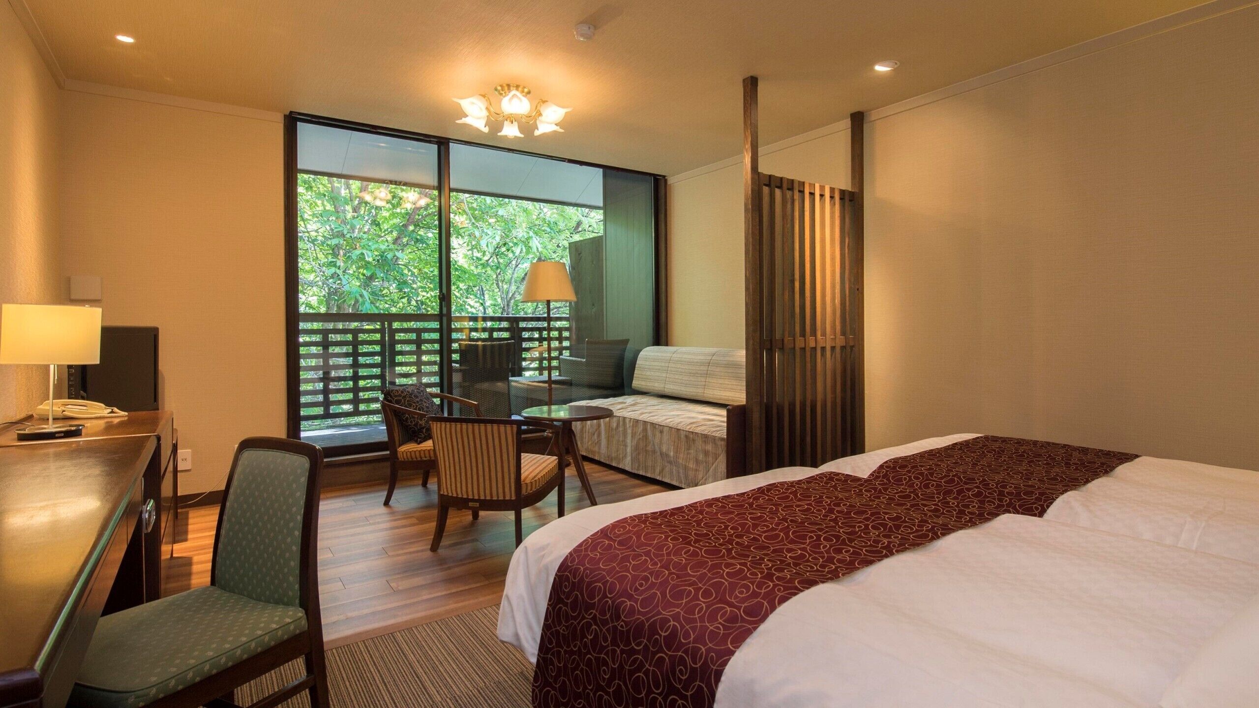 Ein Hotelzimmer mit Bett, Schreibtisch mit Lampe, Stuhl, Balkon mit Tisch und Stühlen sowie einem großen Fenster.