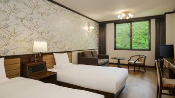 A hotel room with two beds, a TV, a desk, and a window with a view of greenery.