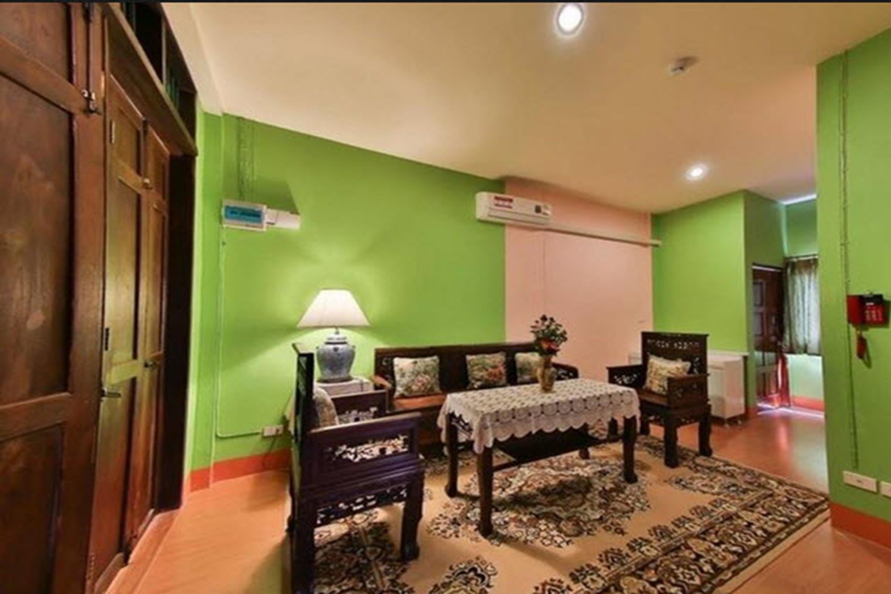 A room with a green wall, a wooden door, a table with a lace tablecloth, a lamp, and a patterned rug.