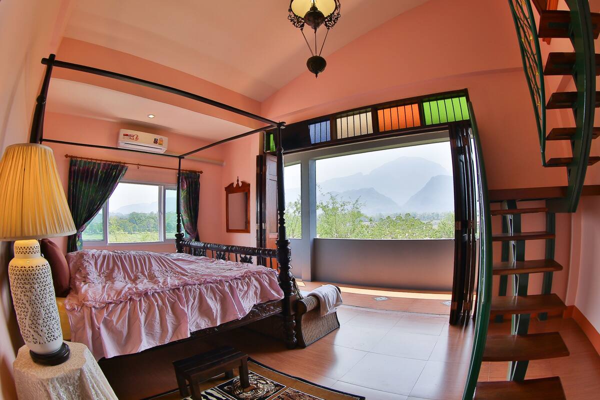 A four-poster bed with a view of mountains through large windows, a staircase, and a table with a lamp.