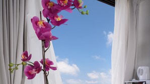 A white table with two white cups, a white vase with pink orchids, and a view of the sea and buildings through a window with white curtains. - Beach View Apartments And Studio (Tel Aviv)