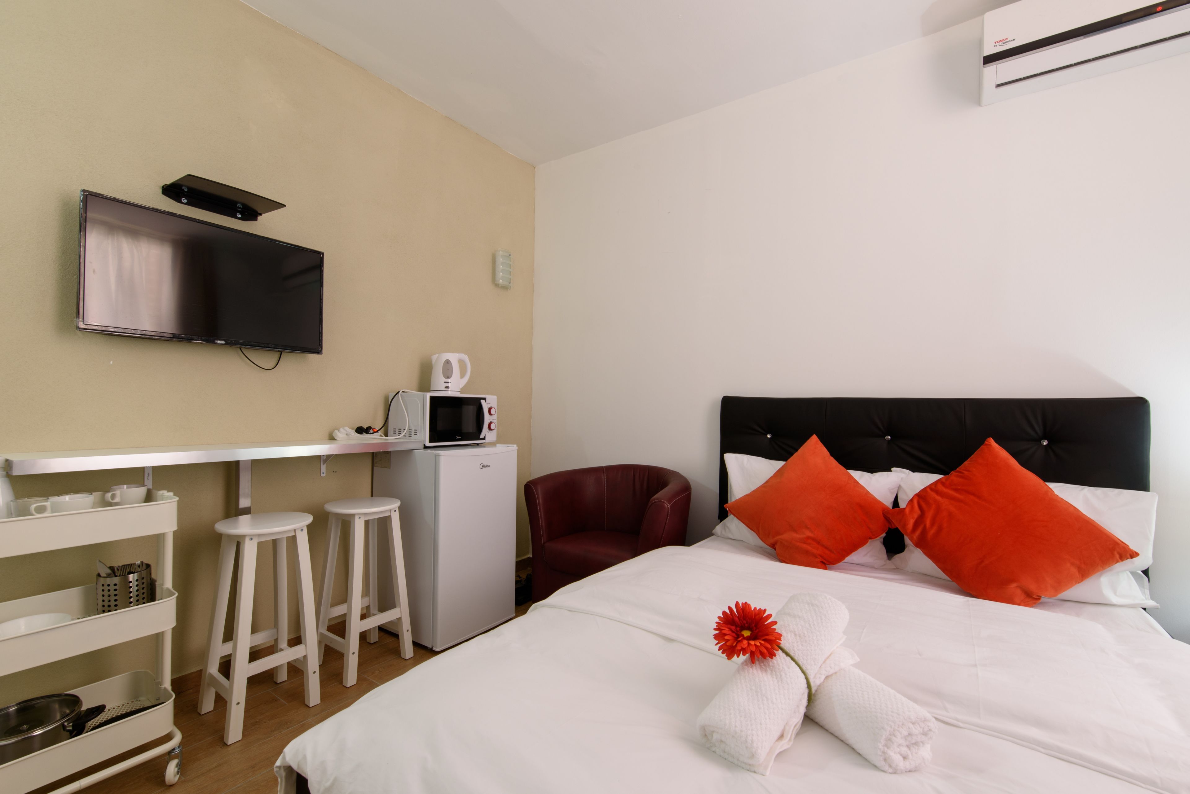 A hotel room with a bed, a TV mounted on the wall, a small kitchenette, and a desk with stools.
