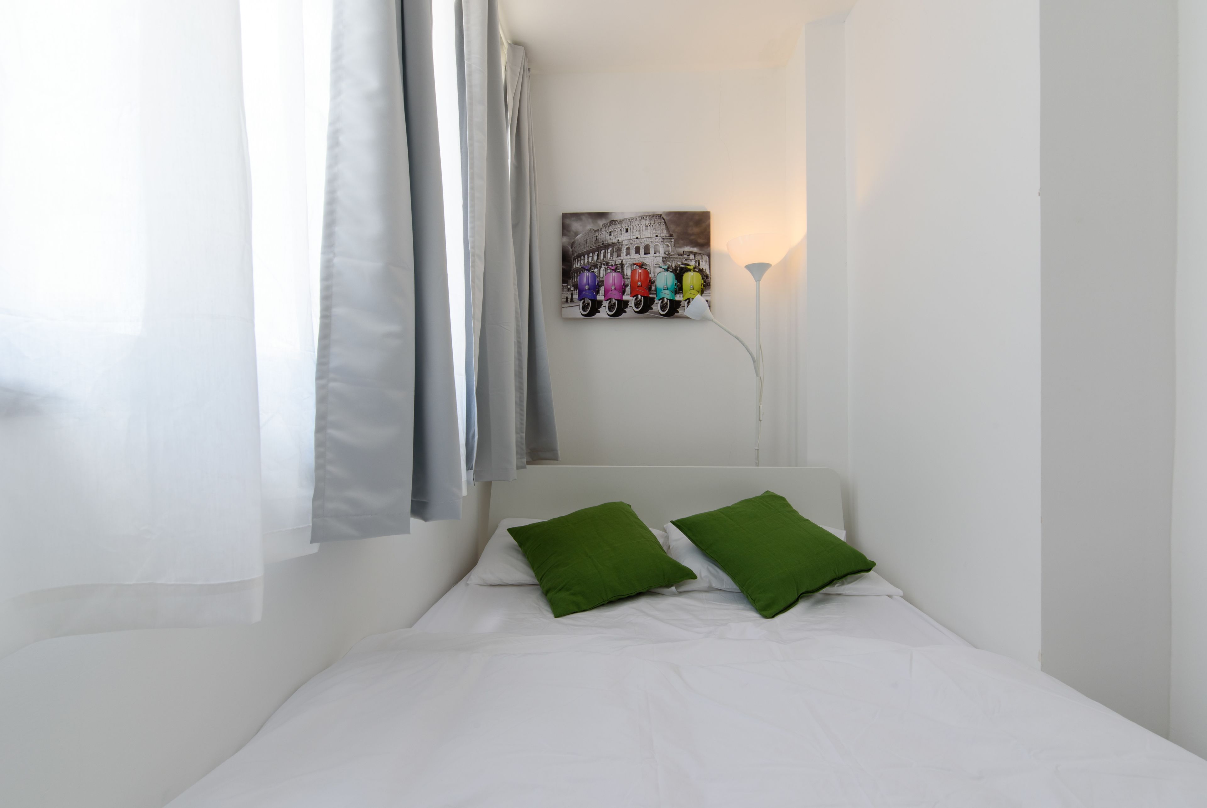 A small, neatly made bed with white linens and two green pillows, a wall-mounted lamp, and a colorful artwork of the Colosseum.