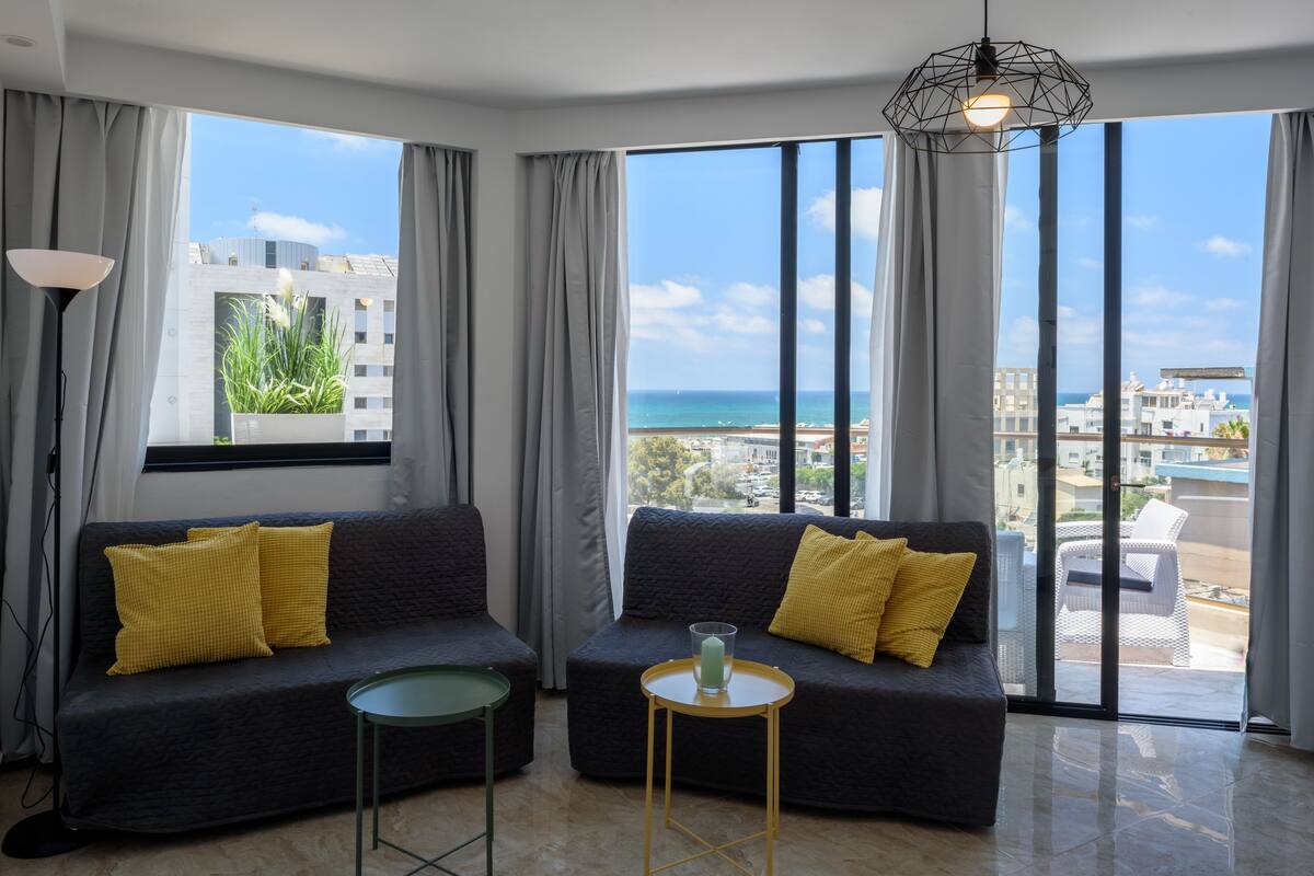 A modern living room with a sofa and a small table, large windows with a view of the sea and buildings, and a hanging light fixture.