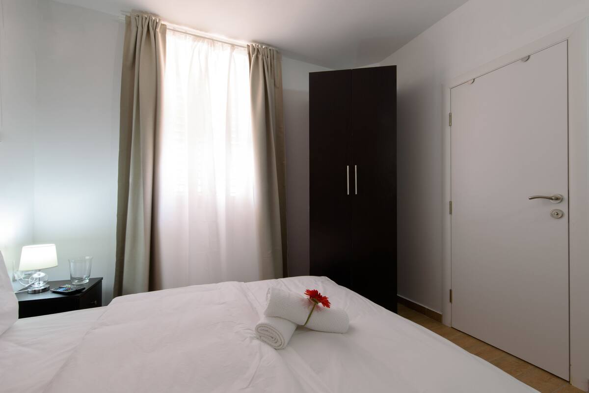 A hotel room with a bed, a nightstand with a lamp, a glass of water, and a dark wardrobe.