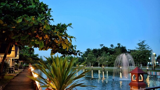 Une scène paisible, avec une fontaine, un sentier en bois et un petit kiosque au bord d’un plan d’eau calme, le tout entouré d’une végétation luxuriante.