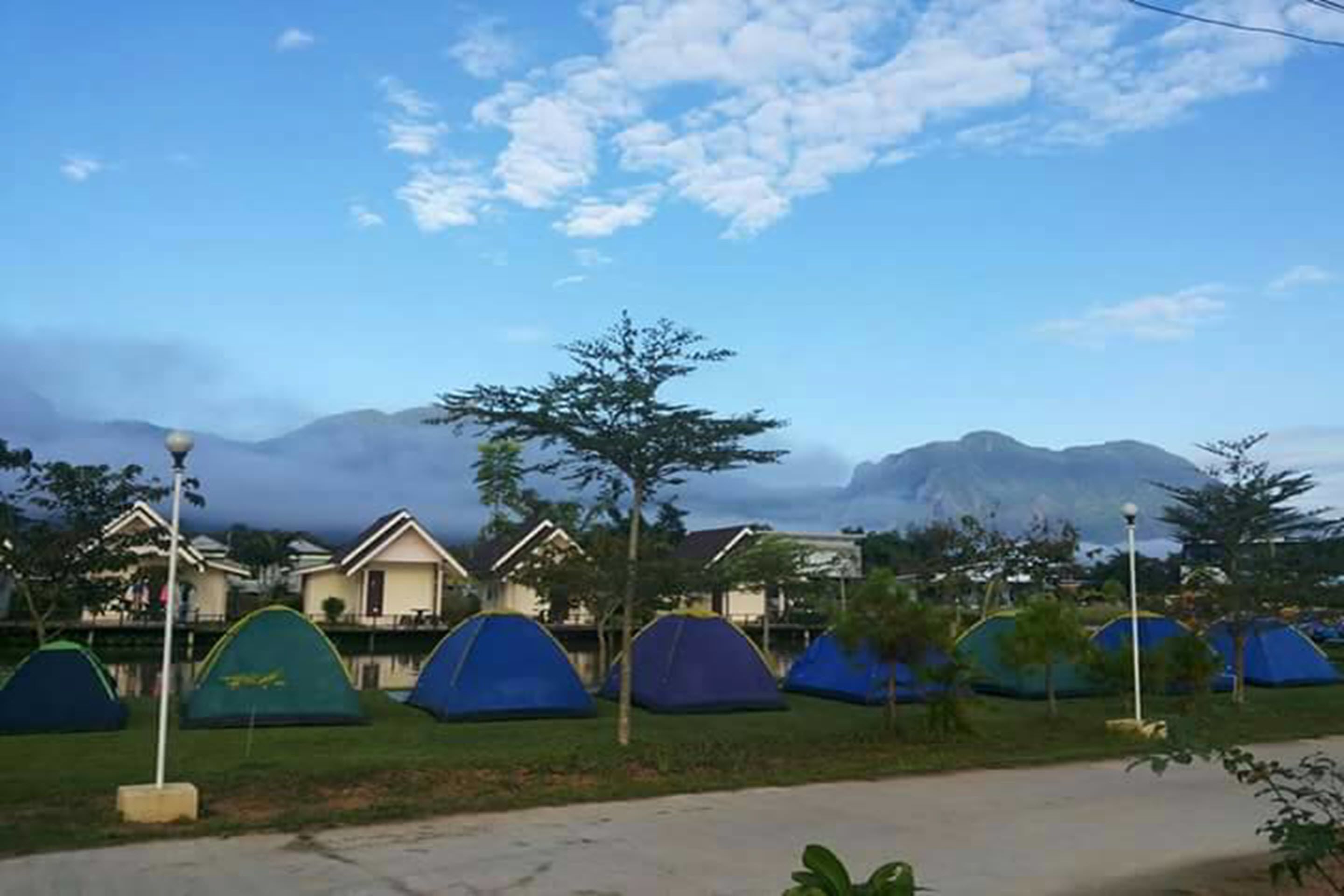 Una hilera de tiendas de campaña en un camping, con casas al fondo y montañas a lo lejos.
