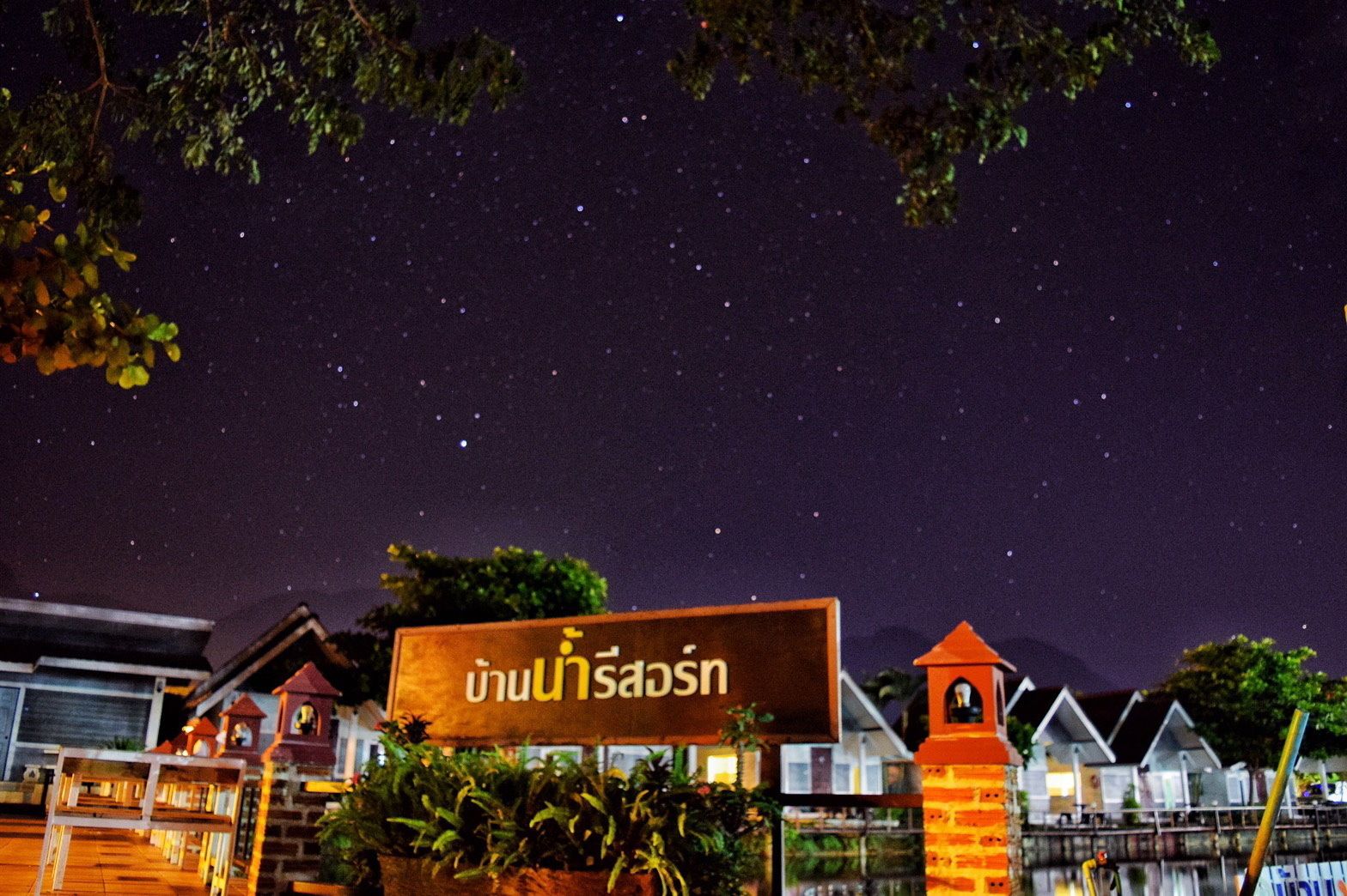 Une scène nocturne avec un ciel étoilé, une enseigne avec du texte en thaï et des exemples d’architecture traditionnelle thaïlandaise.