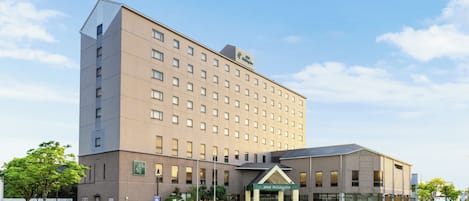 A multi-story hotel with a flat facade, a prominent entrance with a green awning, and a parking lot with cars.