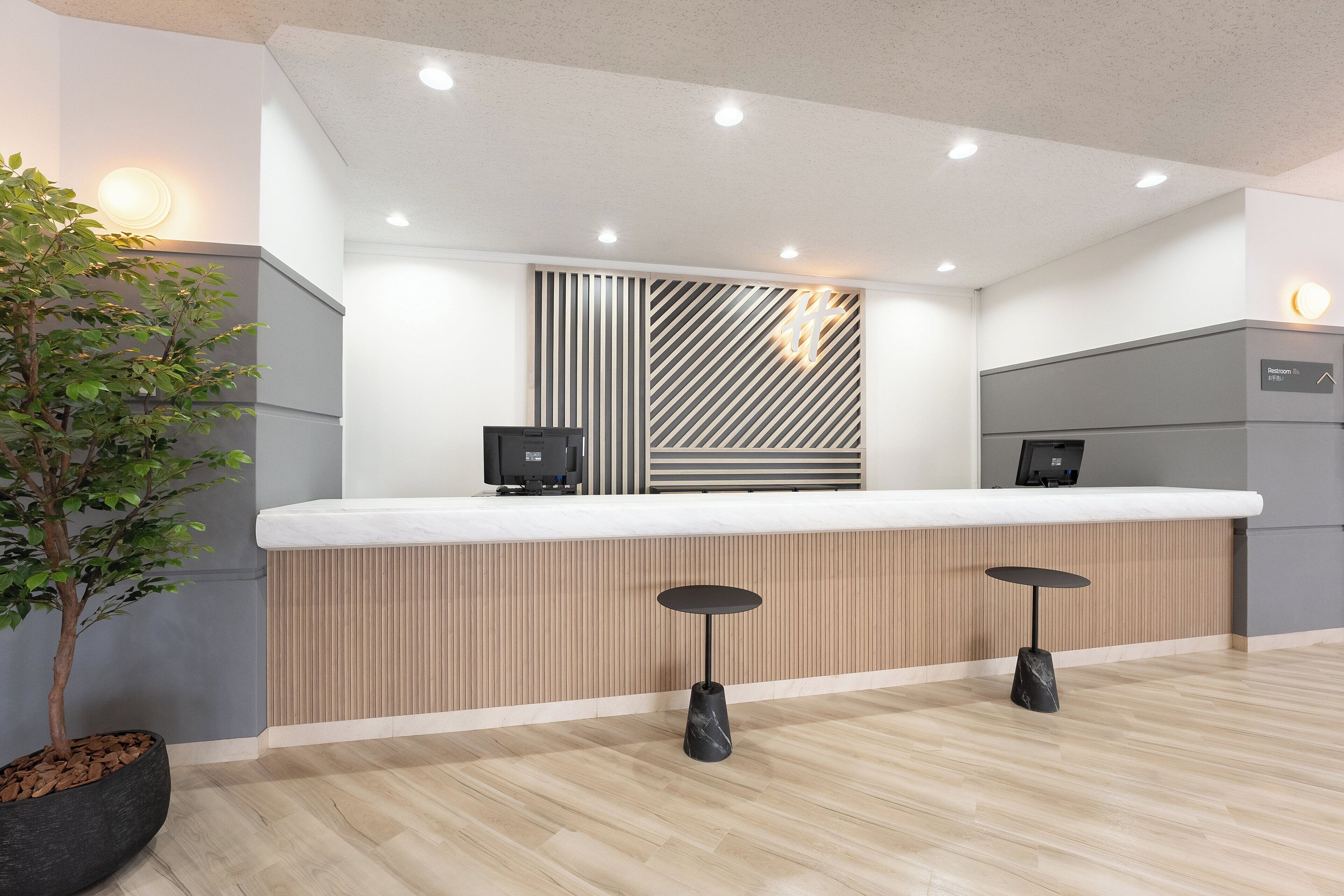 Un mostrador de recepción con diseño de paneles de madera, dos taburetes de bar, una planta en maceta y un cartel que indica los baños.