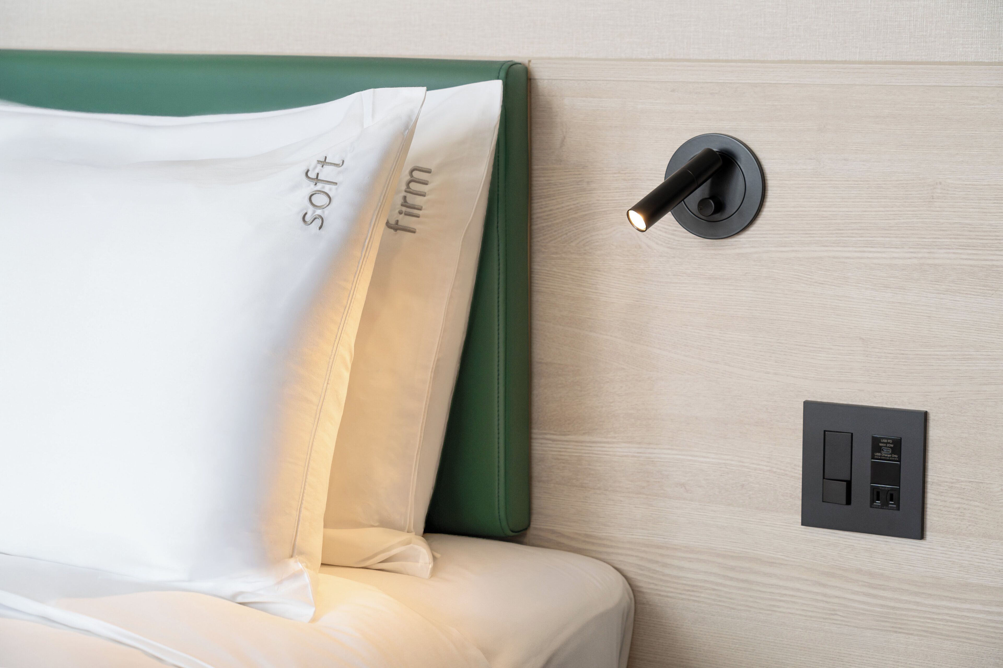 Una cama con cabecera verde, un almohadón blanco con las palabras 'soft' y 'firm', e interruptor junto a la cama con puerto USB.