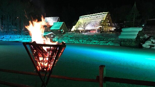 Cena noturna com fogueira em cesto de metal, chão coberto de neve e edifícios iluminados ao fundo.