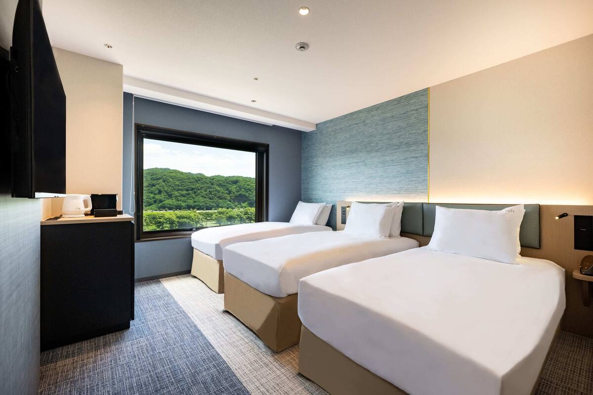 A hotel room with two beds, a desk, and a large window offering a view of greenery.