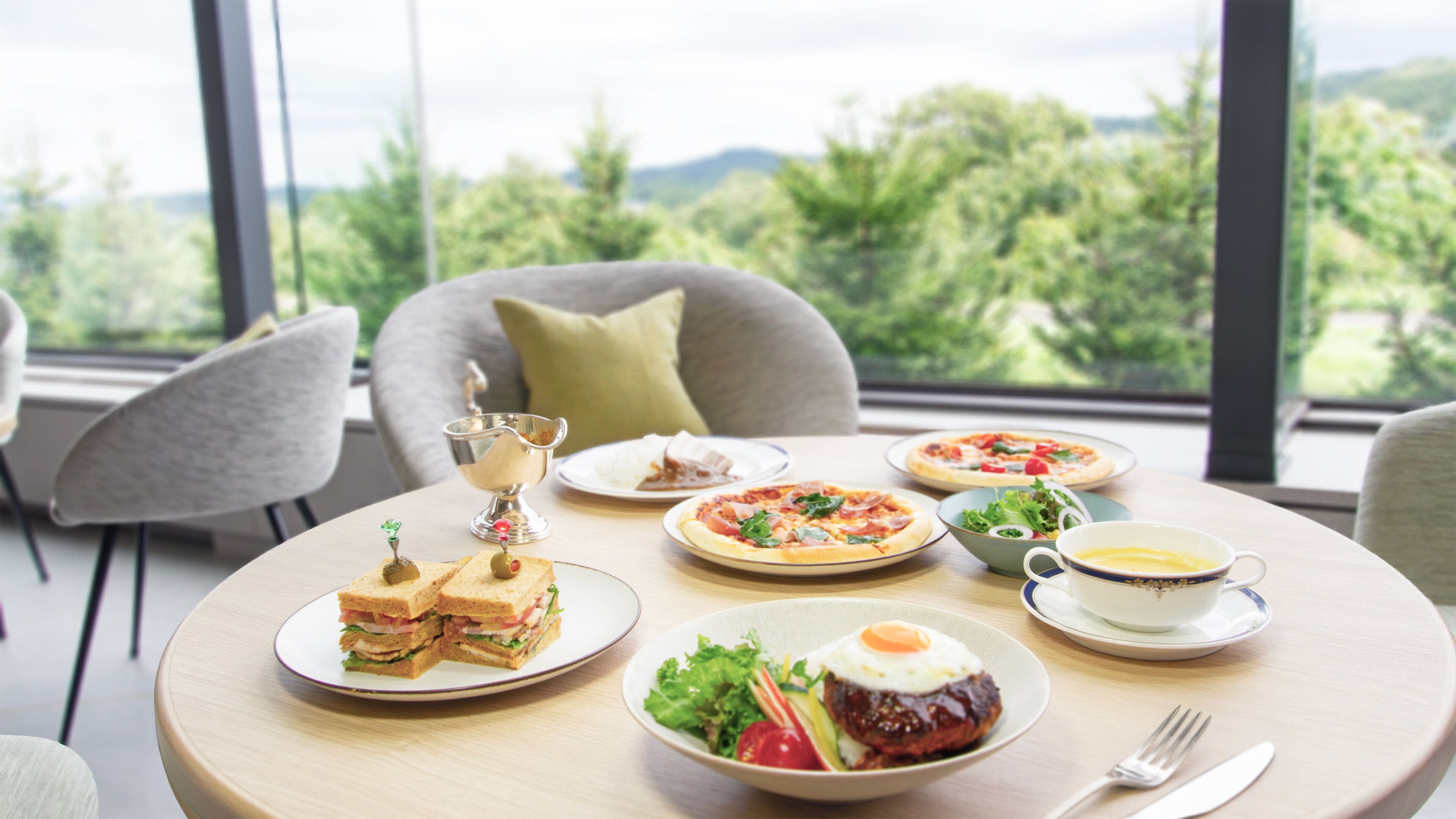 A round dining table with a variety of dishes including sandwiches, pizza, salad, and a cup of coffee. The table is set in a room with large windows offering a view of greenery outside.