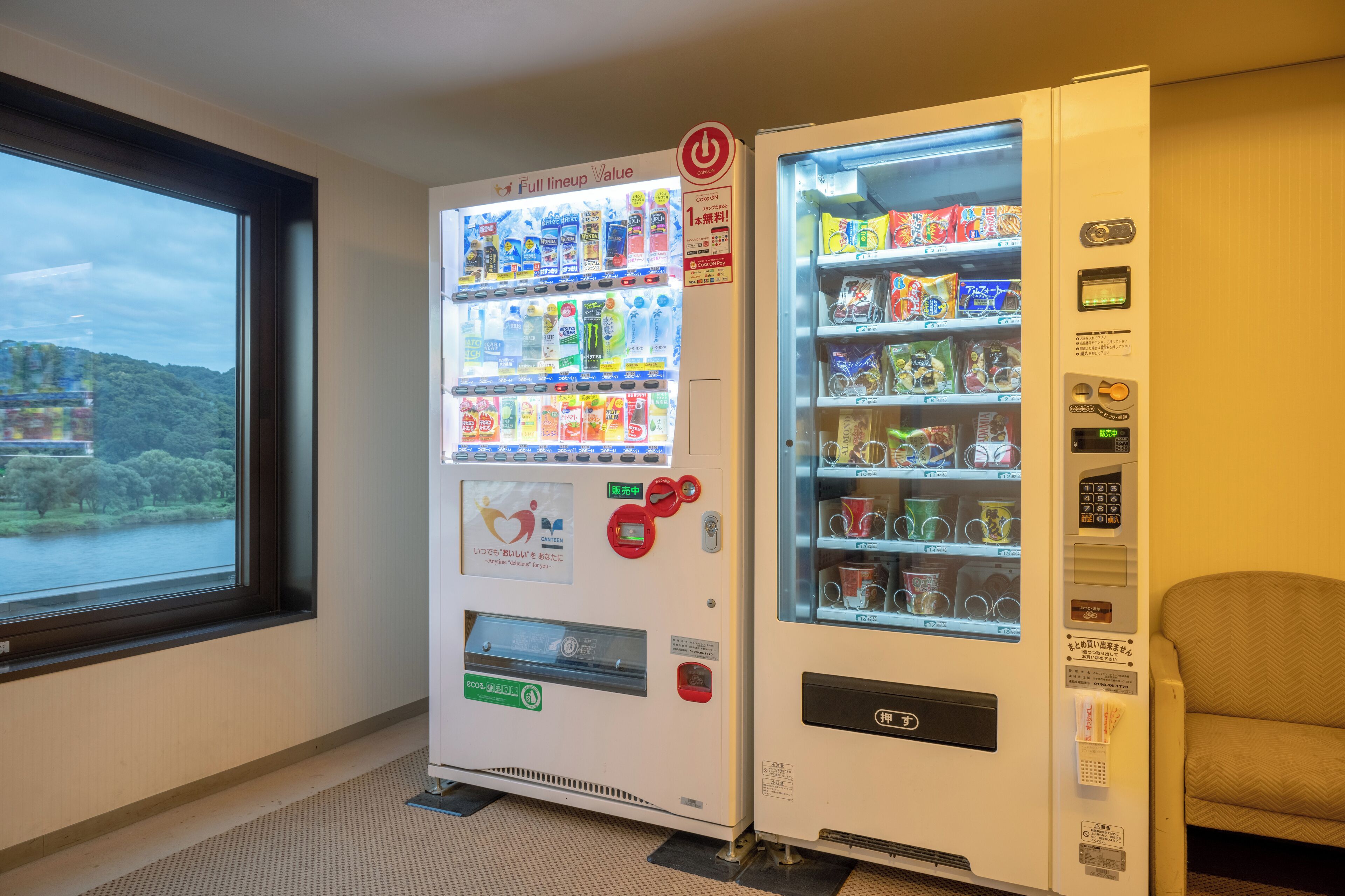 Two vending machines in a room with a window overlooking a natural landscape.