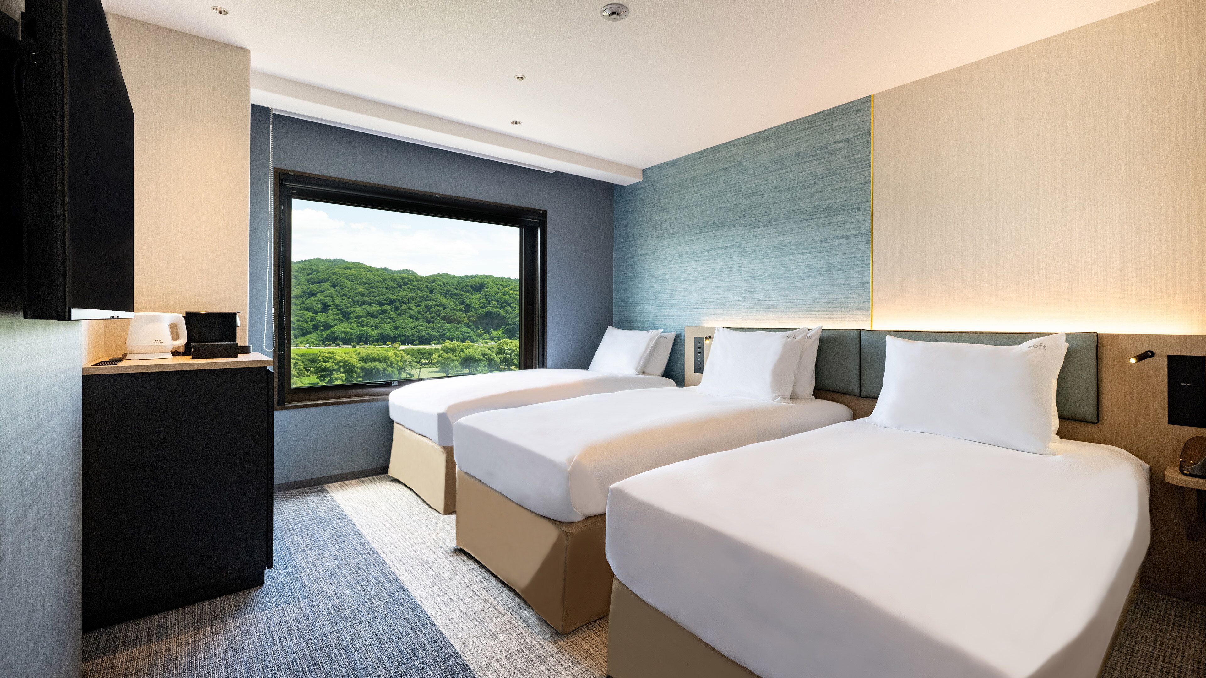 A hotel room with two beds, a desk with a coffee maker, a large window with a view of greenery, and a modern wall design.