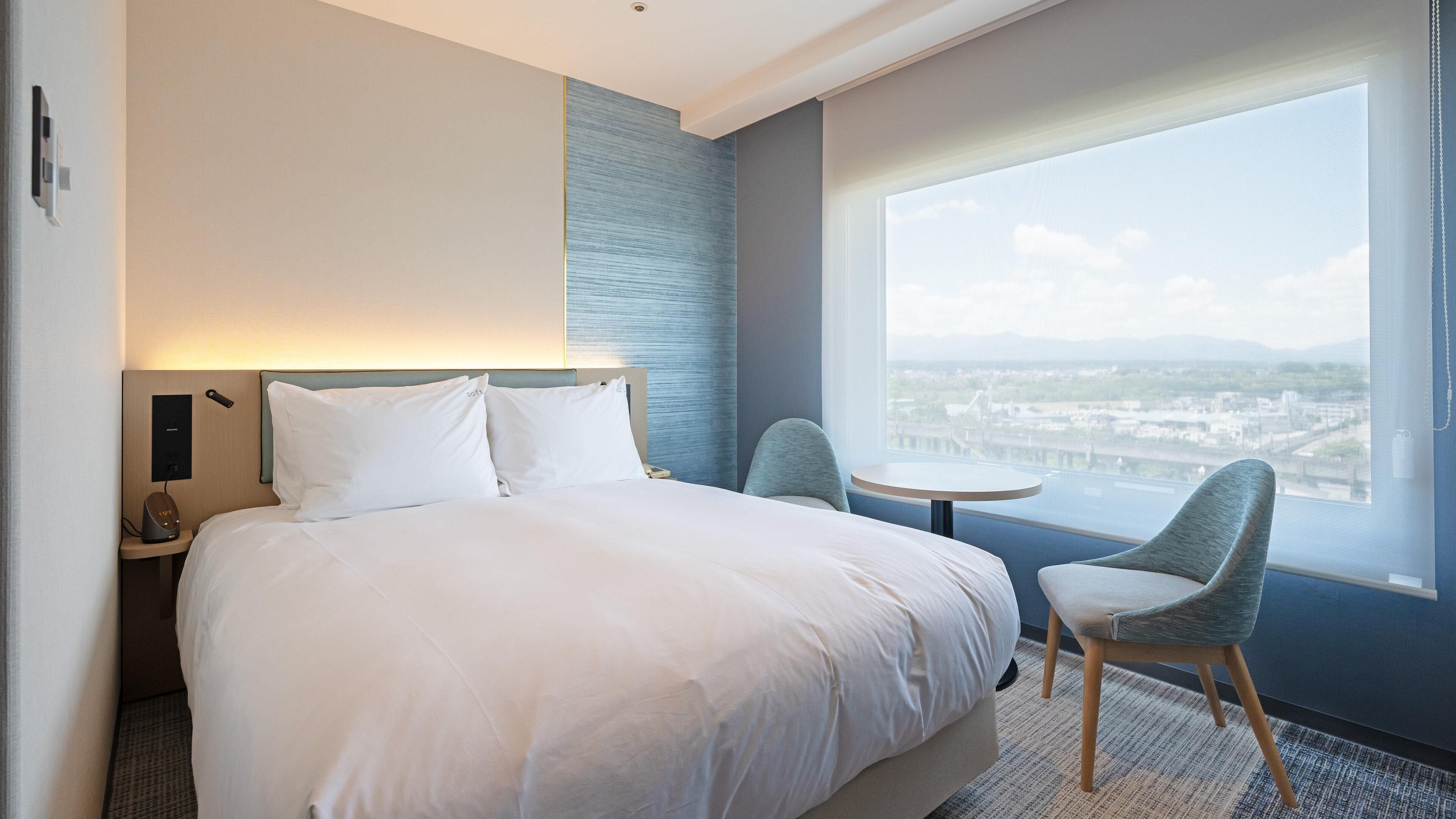 Ein Hotelzimmer mit Bett, Nachttisch, Stuhl und einem großen Fenster mit Blick auf die Stadt.