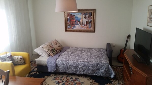 A bedroom with a bed, a chair, a guitar, a TV, and a painting on the wall. - My Home 2 Sea View Apartment (Haifa)
