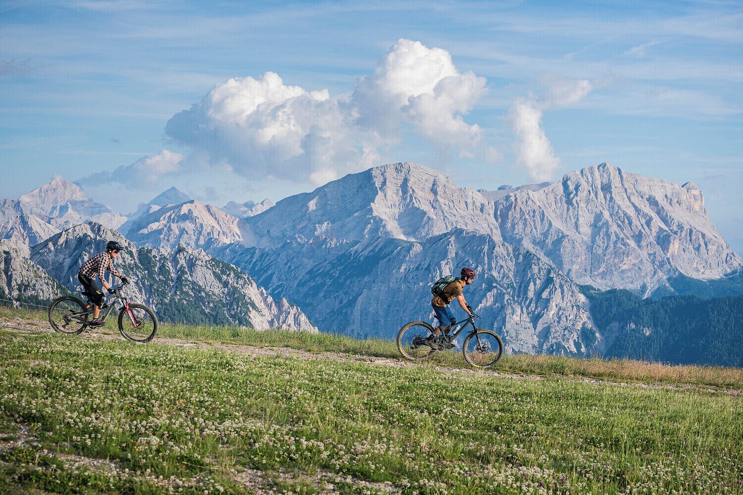 Dos ciclistas de montaña en un sendero, con montañas al fondo.