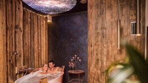 Interior de una cabaña de madera con una bañera grande, una mesita con velas y una lámpara en el techo.