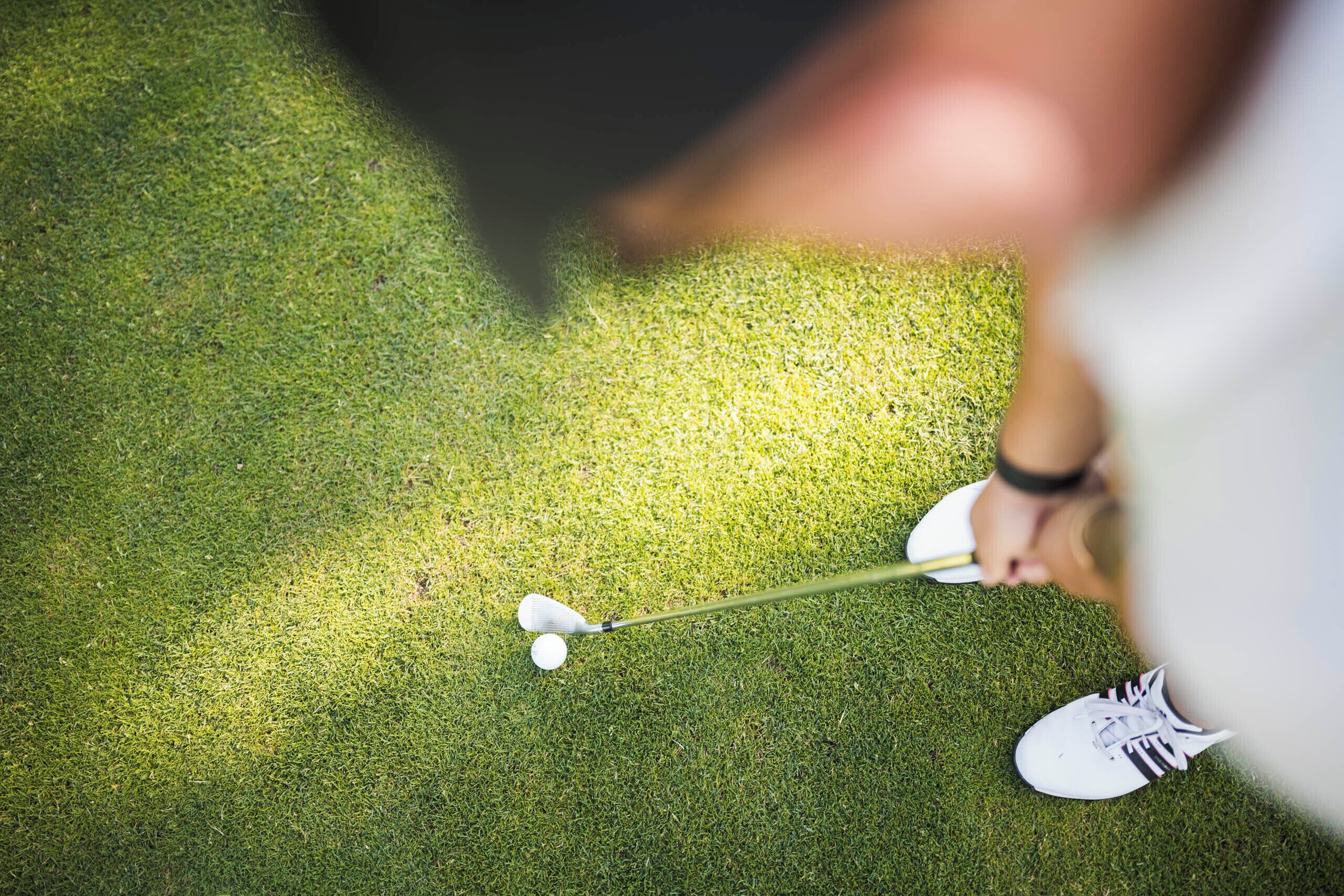 A golf club in motion, a golf ball, and a person's foot on a green surface.