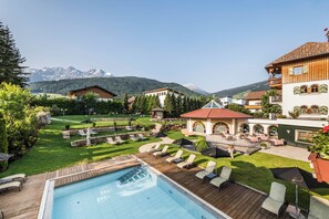 Ein Poolbereich mit Liegestühlen und Sonnenschirmen, ein Garten mit Brunnen und Gebäude mit Blick auf die Berge.