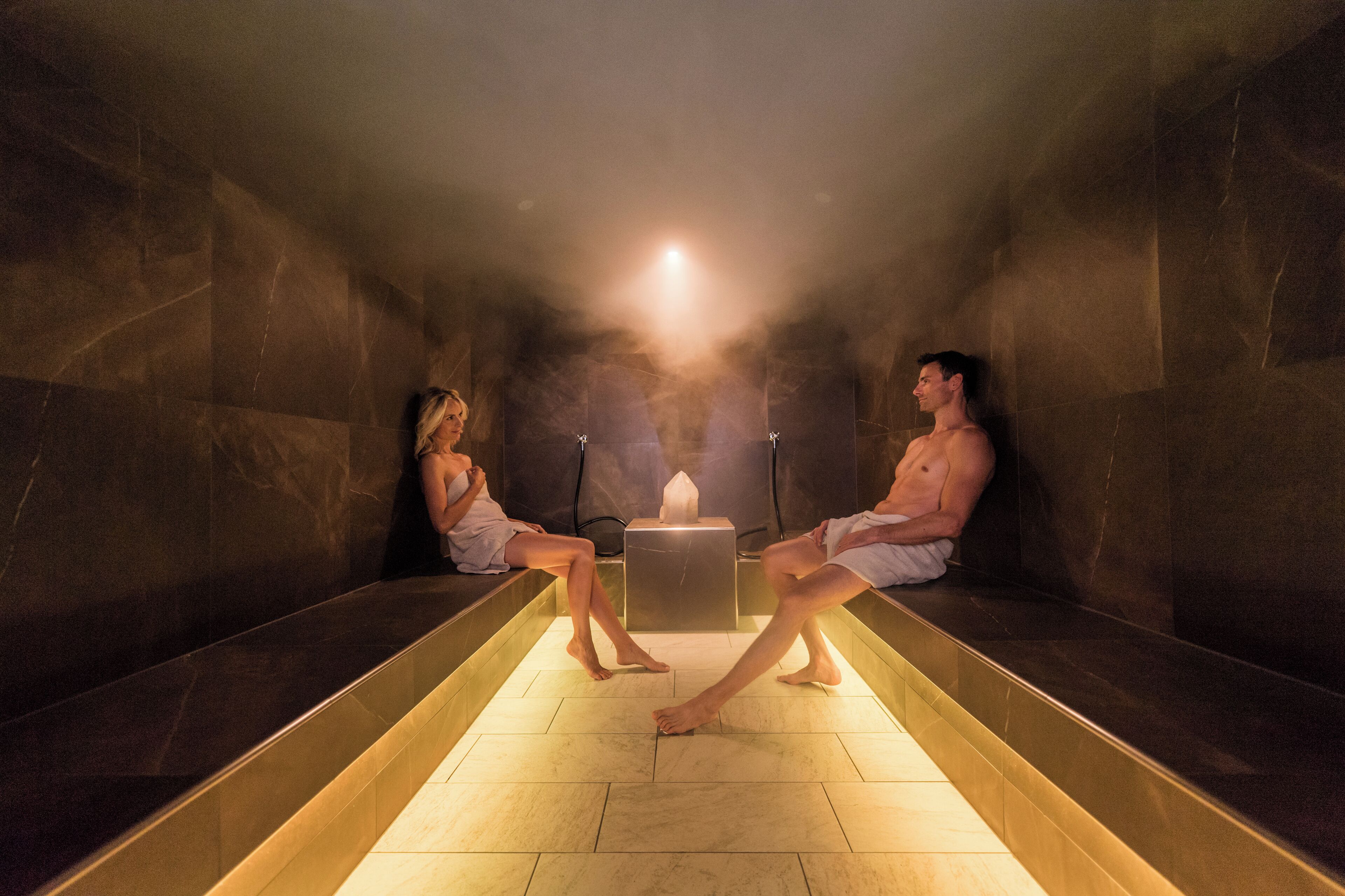 Una sauna con dos personas sentadas en bancos, una fuente de luz central y una pequeña mesa.