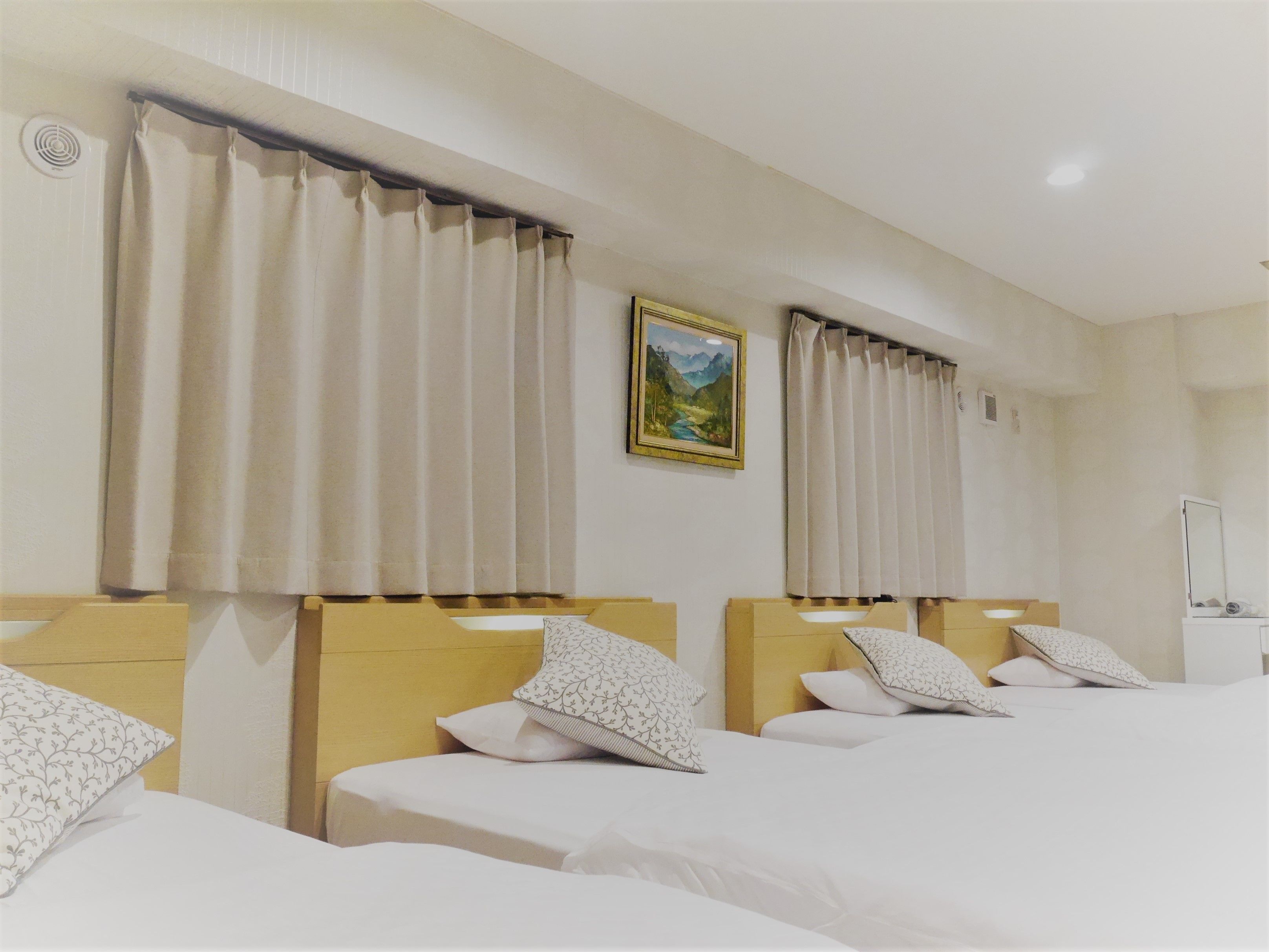 A hotel room with three beds, beige curtains, a painting on the wall, and a bedside table with a lamp.
