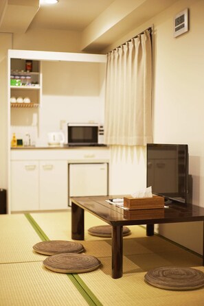 A room with tatami mats, a wooden table, a television, and a kitchen area in the background.