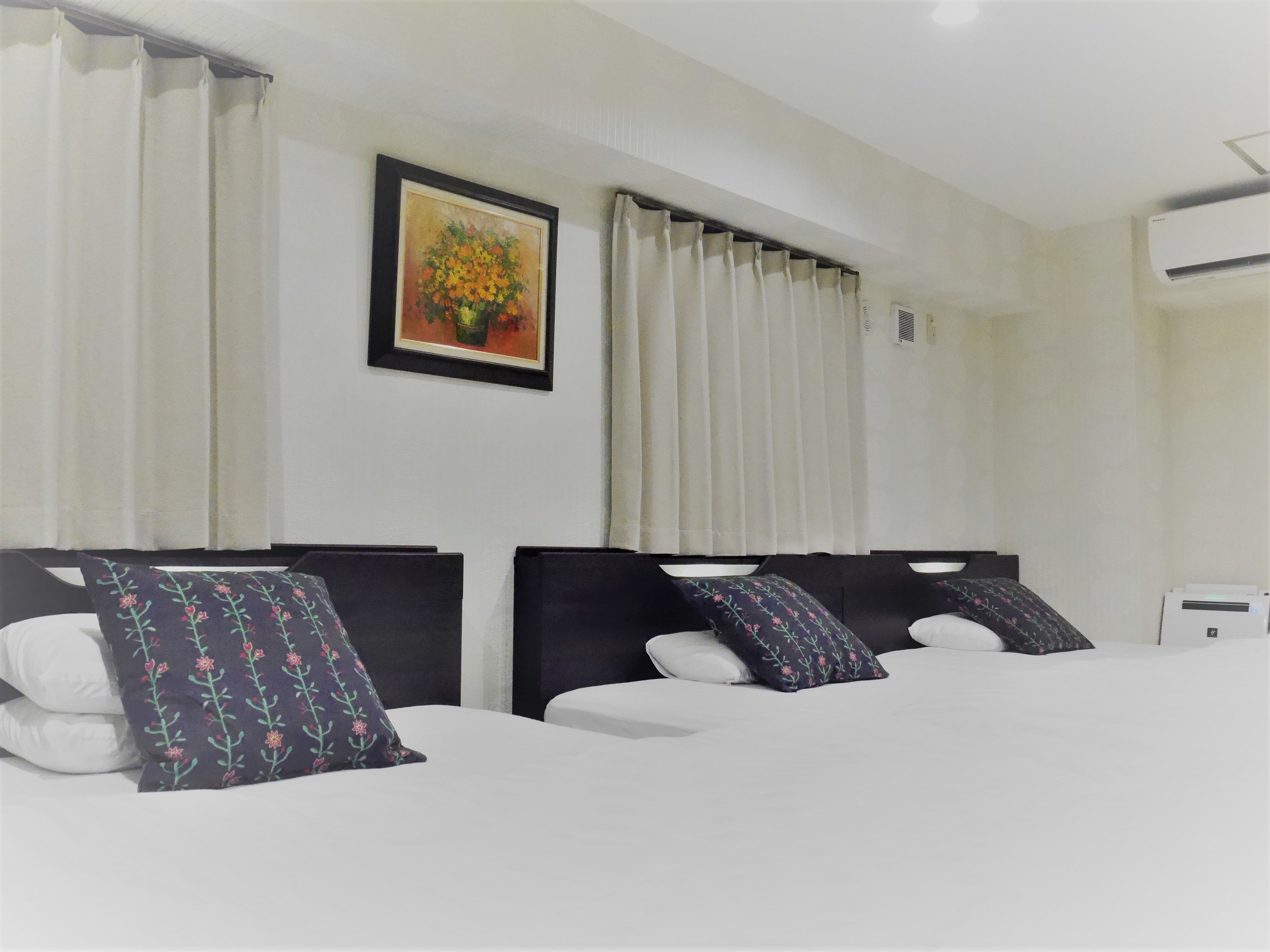 A hotel room with two beds, white bedding, dark headboards, floral-patterned pillows, a framed floral painting on the wall, and an air conditioning unit.