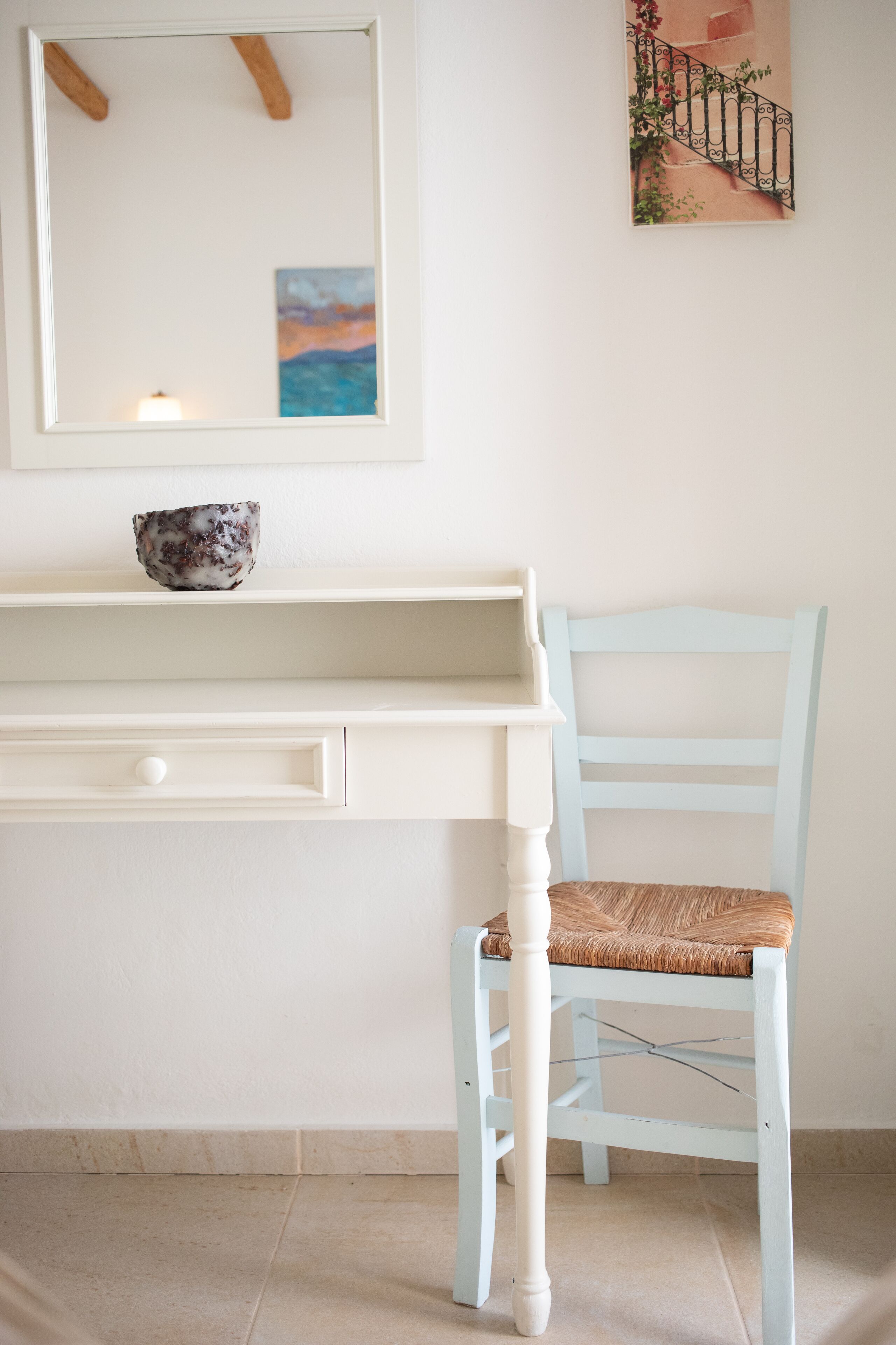 A white desk with a bowl on top, a light blue chair with a woven seat, and a painting of a staircase on the wall.