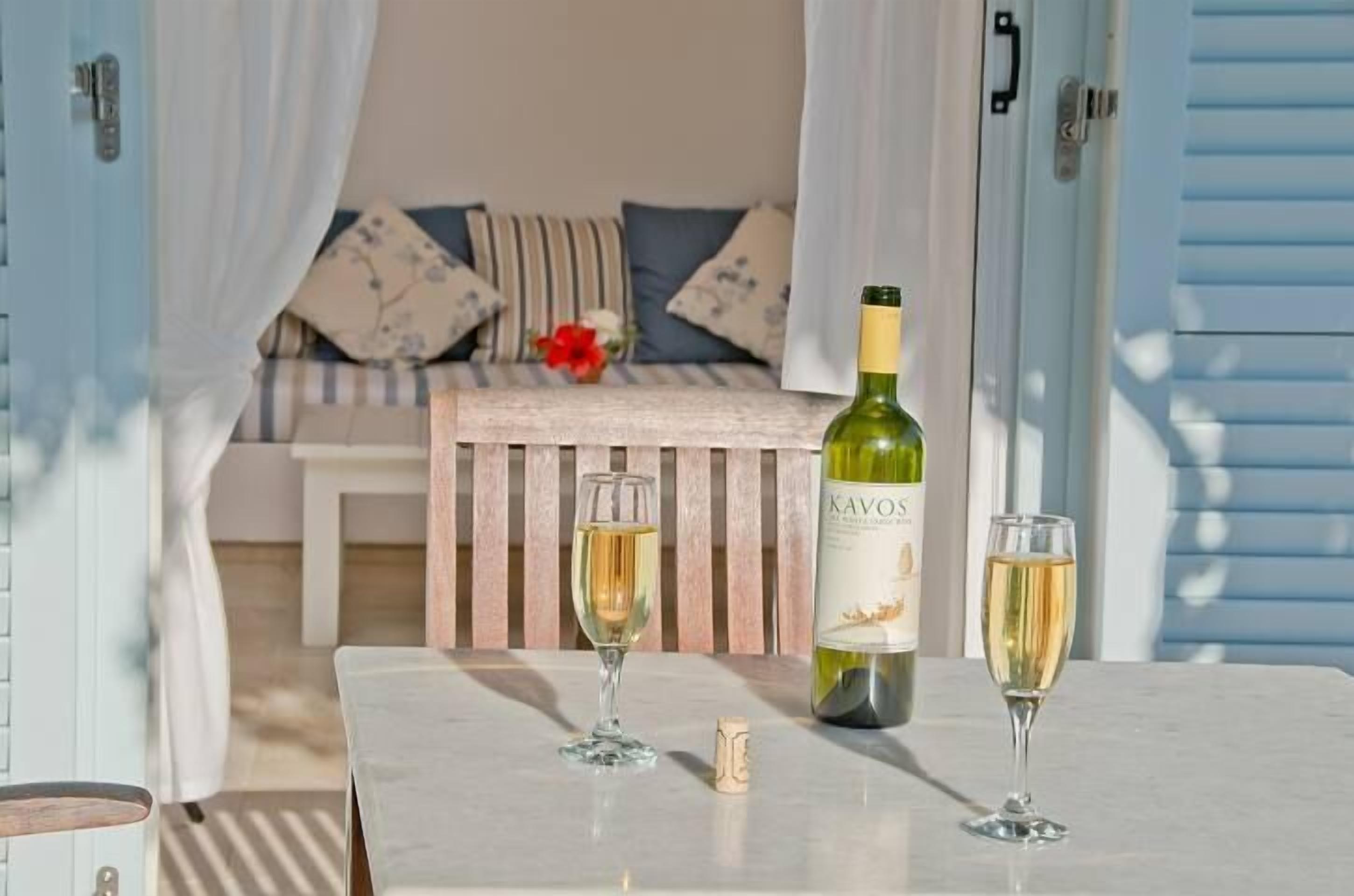 A table with a bottle of Kavos wine and two glasses, a cork, and a striped bench with pillows in a room with light blue shutters.