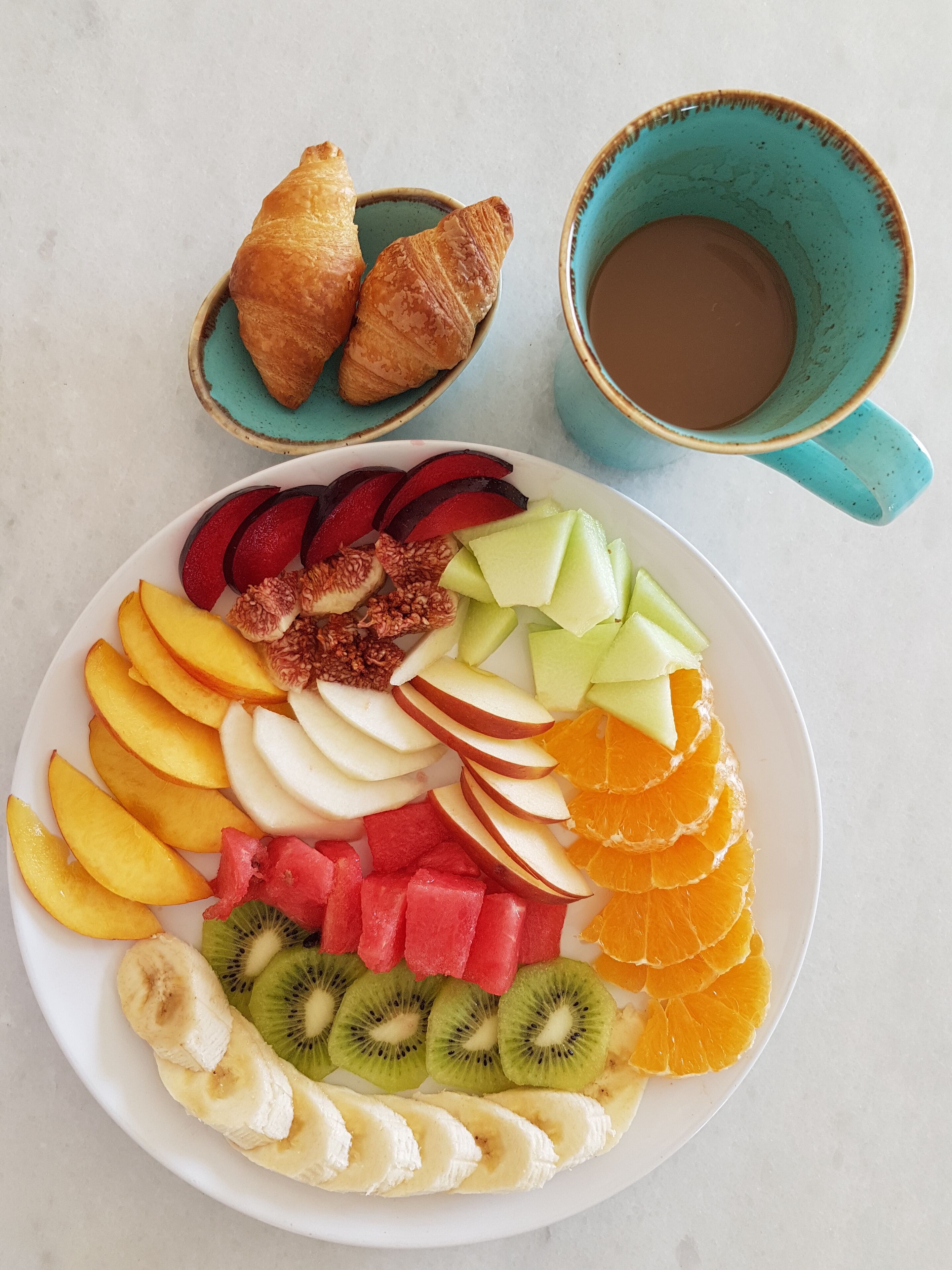 Une assiette de fruits variés et un croissant, une tasse de café et un petit bol de croissants.