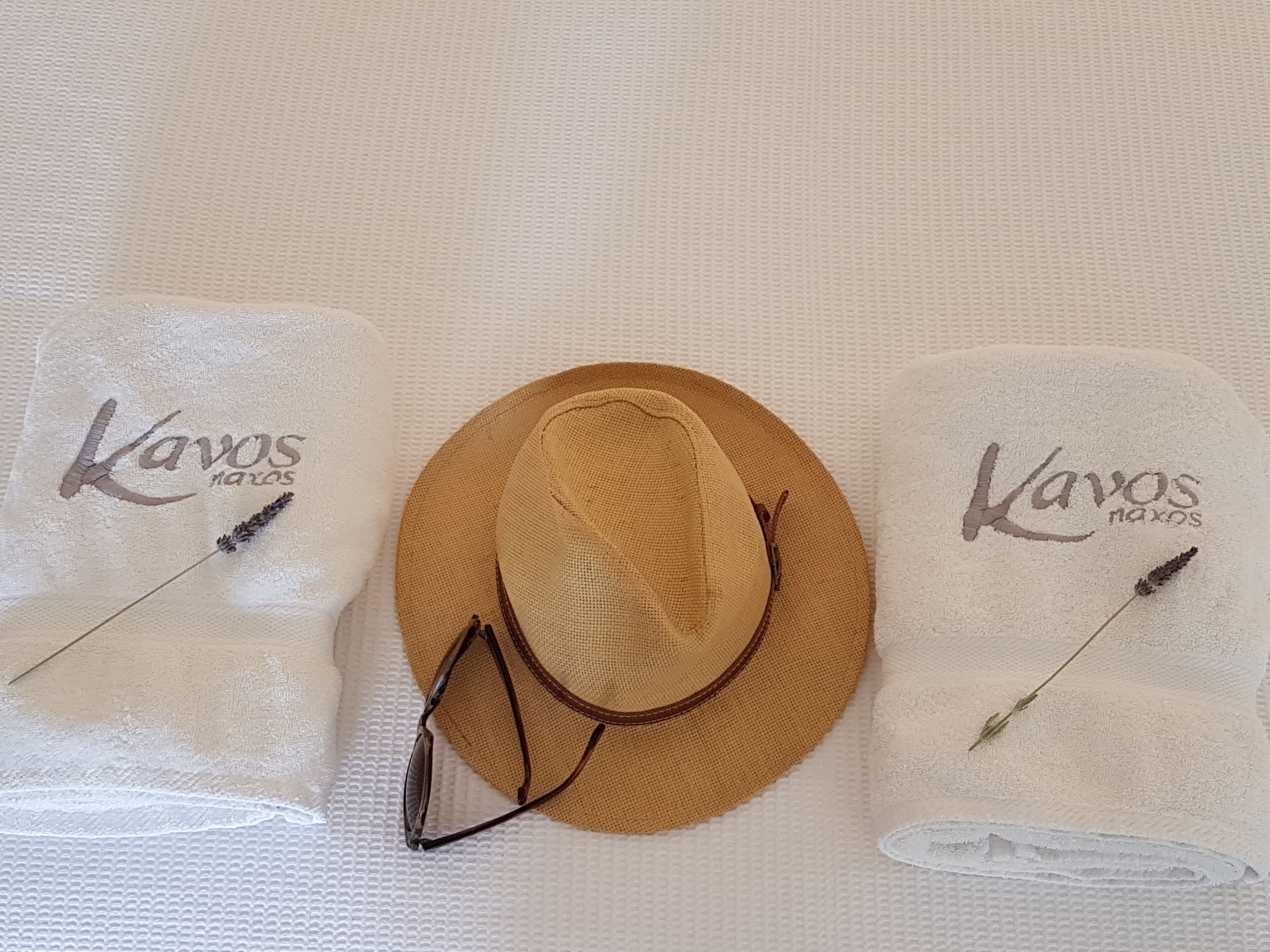 En hatt och ett par solglasögon på en vit, strukturerad yta, tillsammans med texten ”Kavos maxos” och en torkad lavendelkvist.