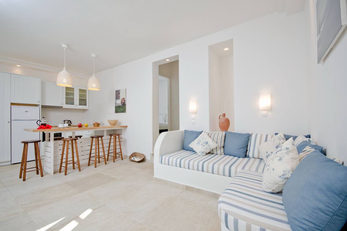 Cocina moderna con armarios blancos, encimera de madera y taburetes de bar. Hay un sofá blanco con cojines a rayas y un jarrón sobre una mesita.