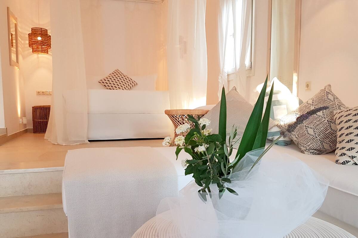 Una habitación moderna y luminosa con un sofá blanco, una mesa redonda blanca con un florero y un ventanal con cortinas translúcidas.