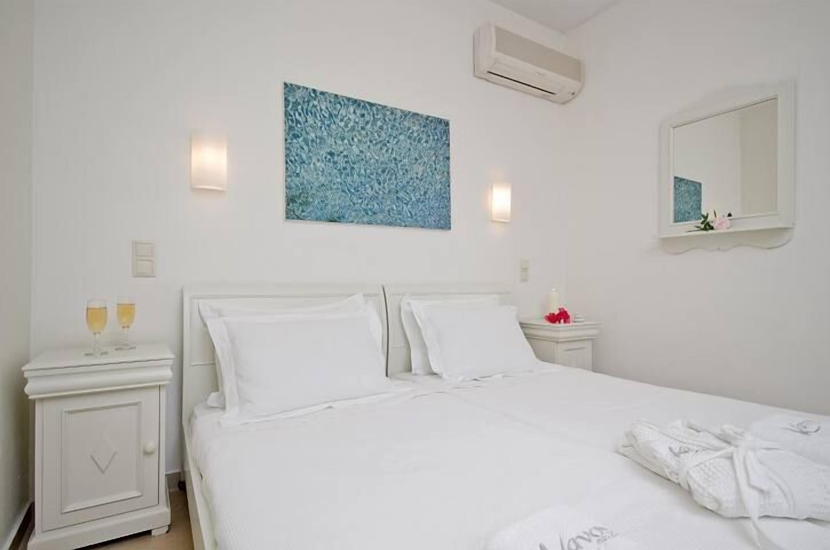 Habitación de hotel con una cama blanca, una mesita de noche con dos copas de vino, un aire acondicionado de pared y un cuadro sobre la cama.