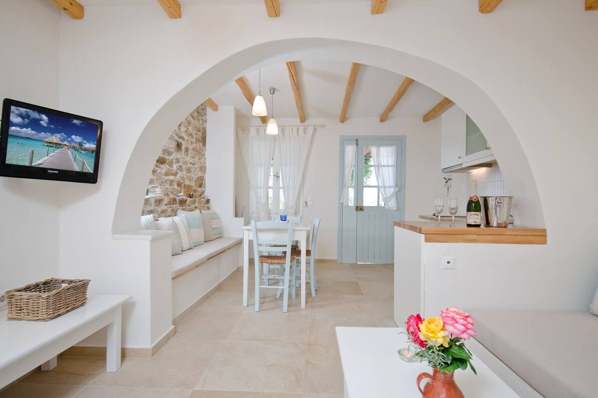 Una habitación acogedora con pared de piedra, un banco blanco, una mesa de comedor con sillas, un televisor montado en la pared y una zona de cocina con mostrador y copas de vino.