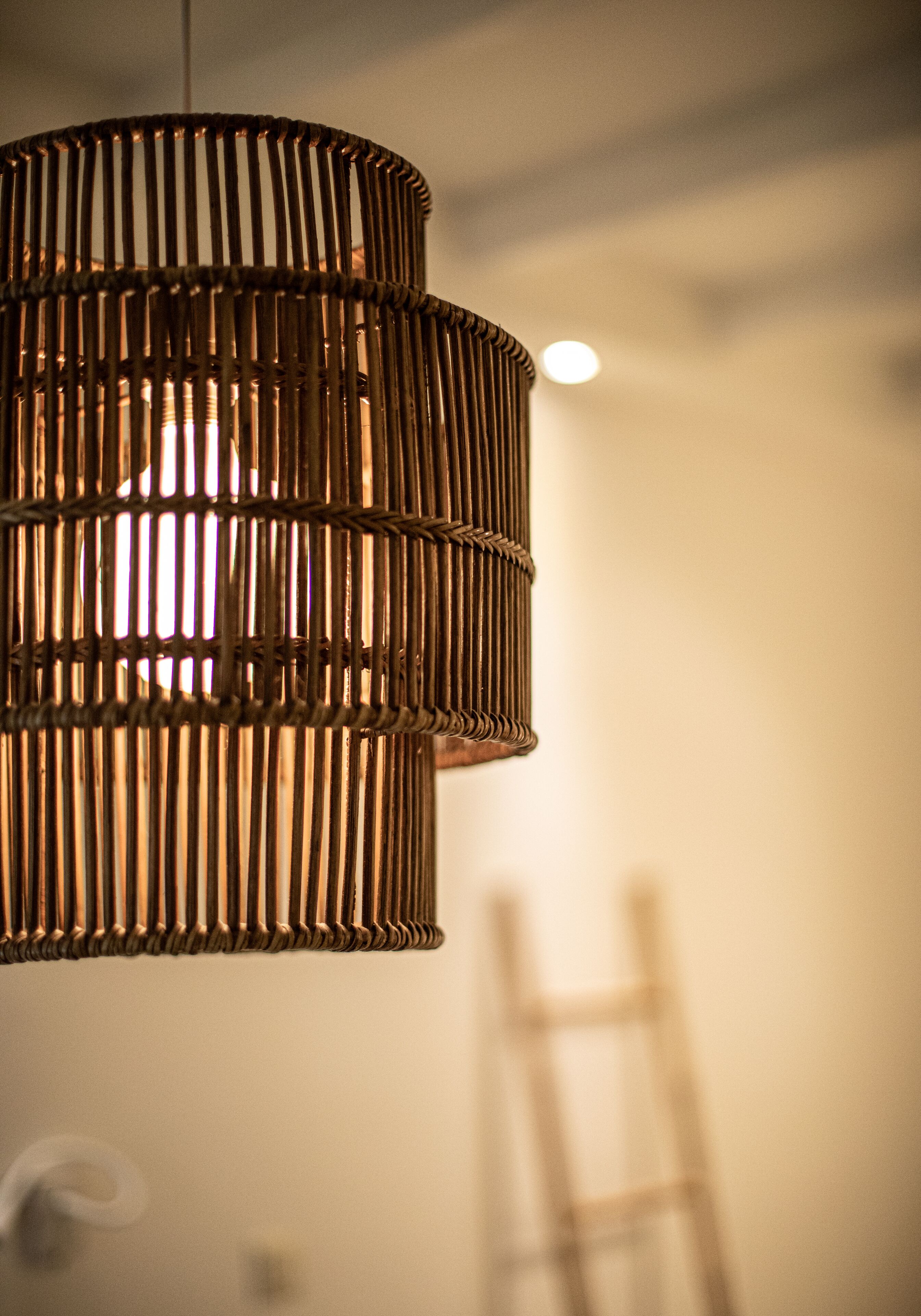 A wicker pendant light fixture with a soft glow, hanging in a room with a ladder in the background.