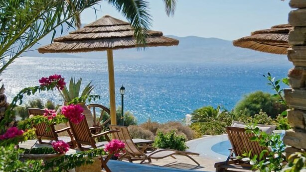 Zona de piscina con sillones de descanso y una sombrilla de paja, con vistas panorámicas del mar y las montañas.