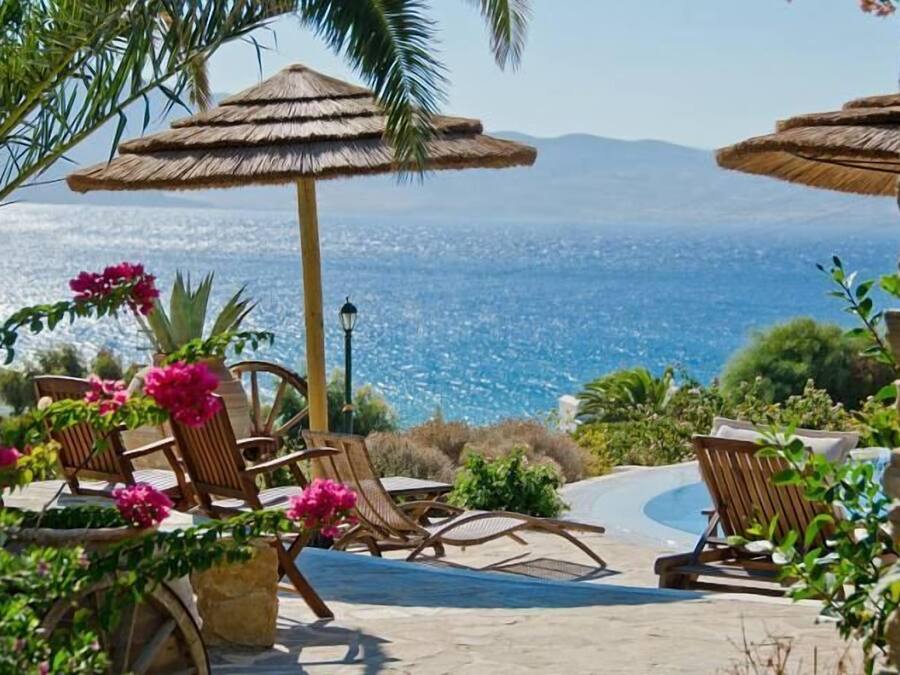 Zona de piscina con sillones de descanso y una sombrilla de paja, con vistas panorámicas del mar y las montañas.