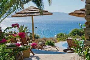 Zona de piscina con sillones de descanso y una sombrilla de paja, con vistas panorámicas del mar y las montañas.