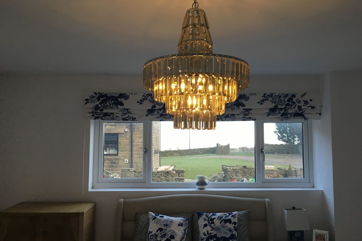 A bedroom with a bed, a chandelier, a window with a view of the outdoors, and a bedside table with a lamp and a picture frame.