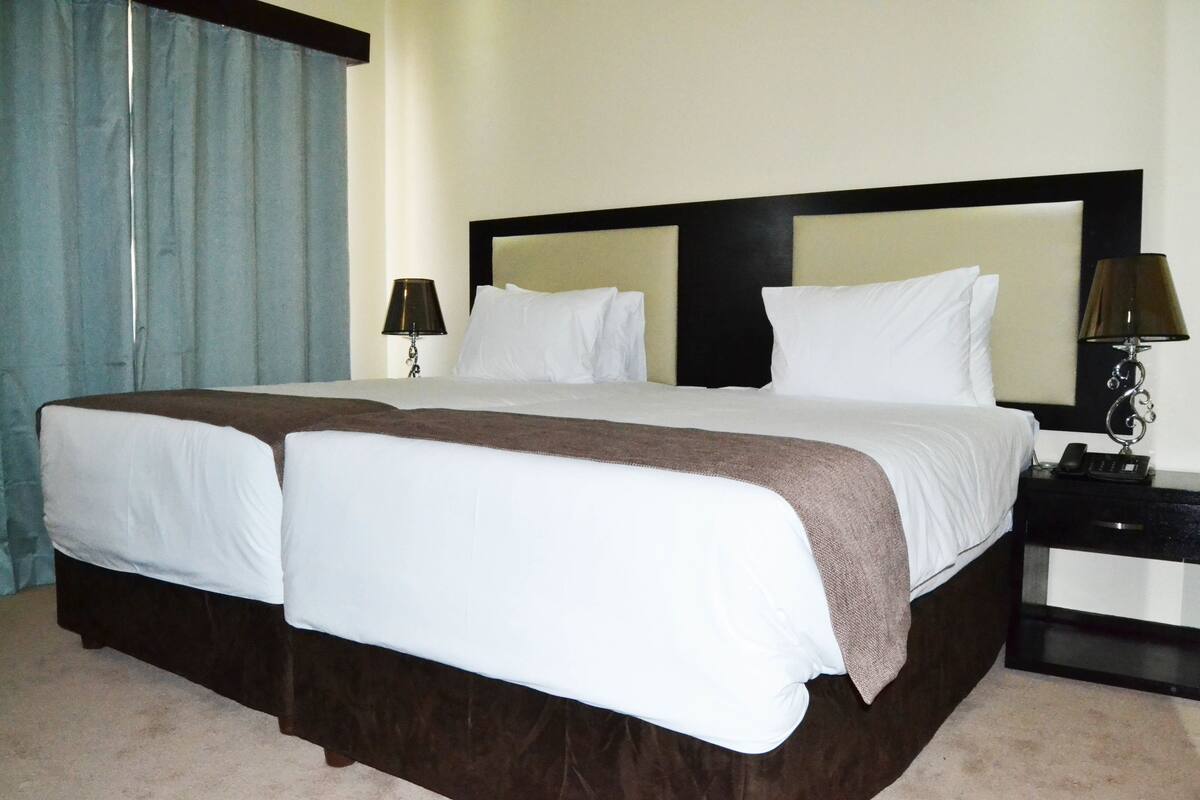 Une chambre d’hôtel avec un grand lit, deux tables de chevet, une petite table et une fenêtre avec des rideaux.