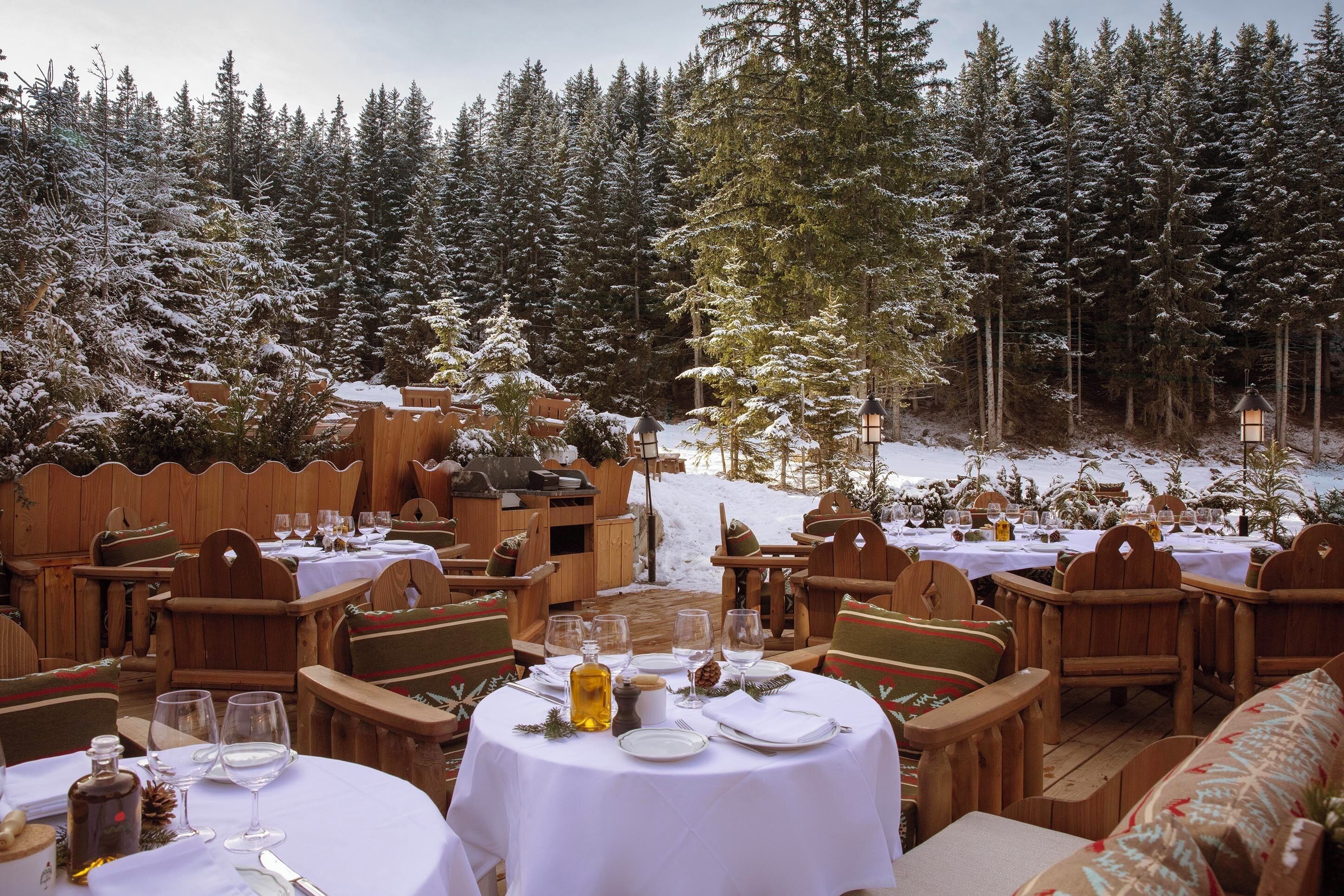Un espace de restauration en plein air, avec des tables rondes dressées pour un repas, entourées de chaises et de bancs en bois, et une forêt enneigée en arrière-plan.