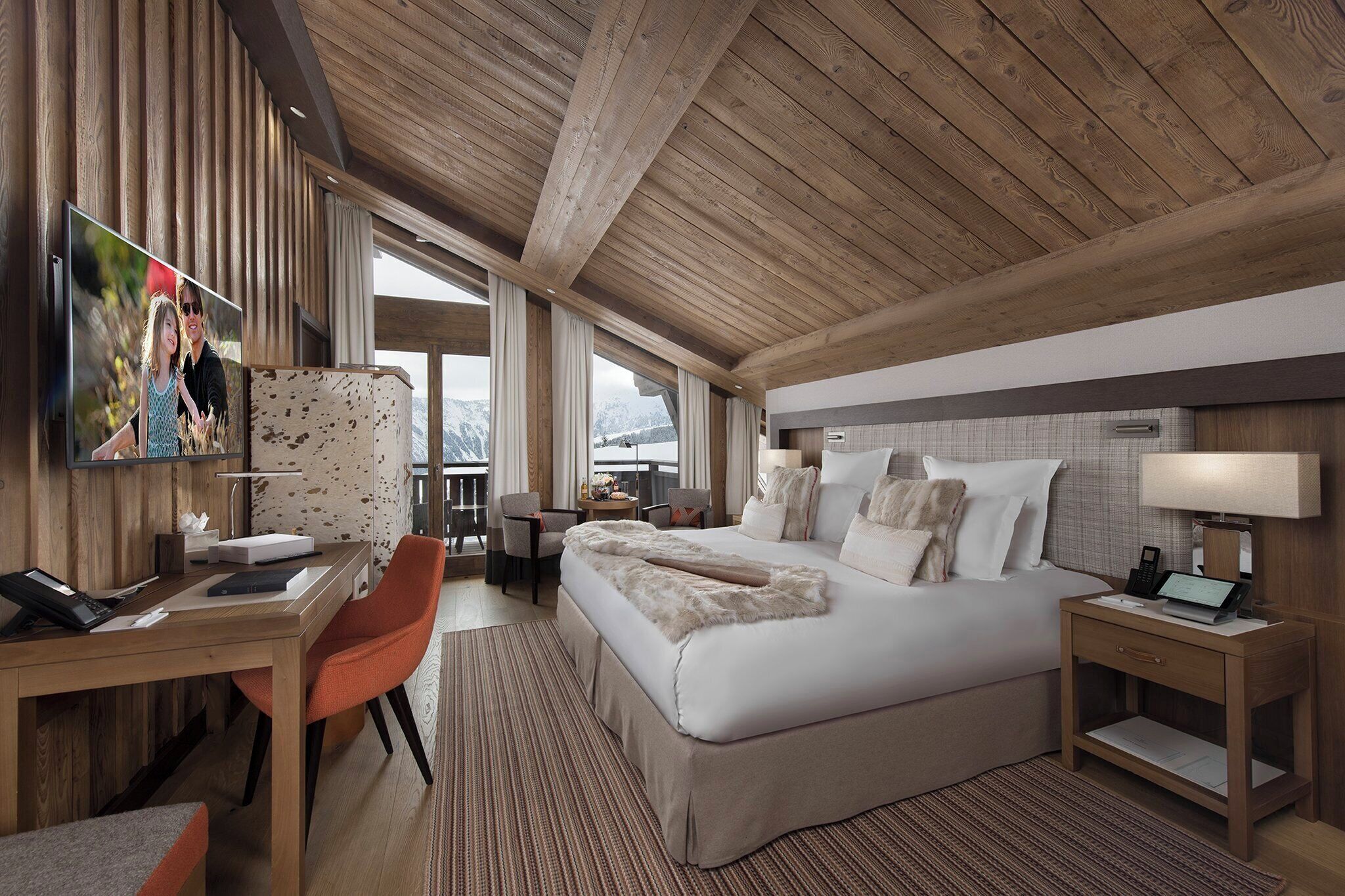 A modern hotel room with a wooden ceiling, a large bed with white bedding, a desk with a chair, a TV mounted on the wall, and a view of mountains through large windows.
