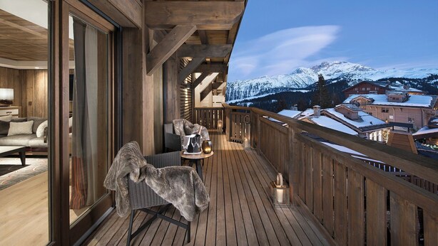 A wooden deck with a view of snow-covered mountains, a cozy seating area with a chair and a table, and a large window.