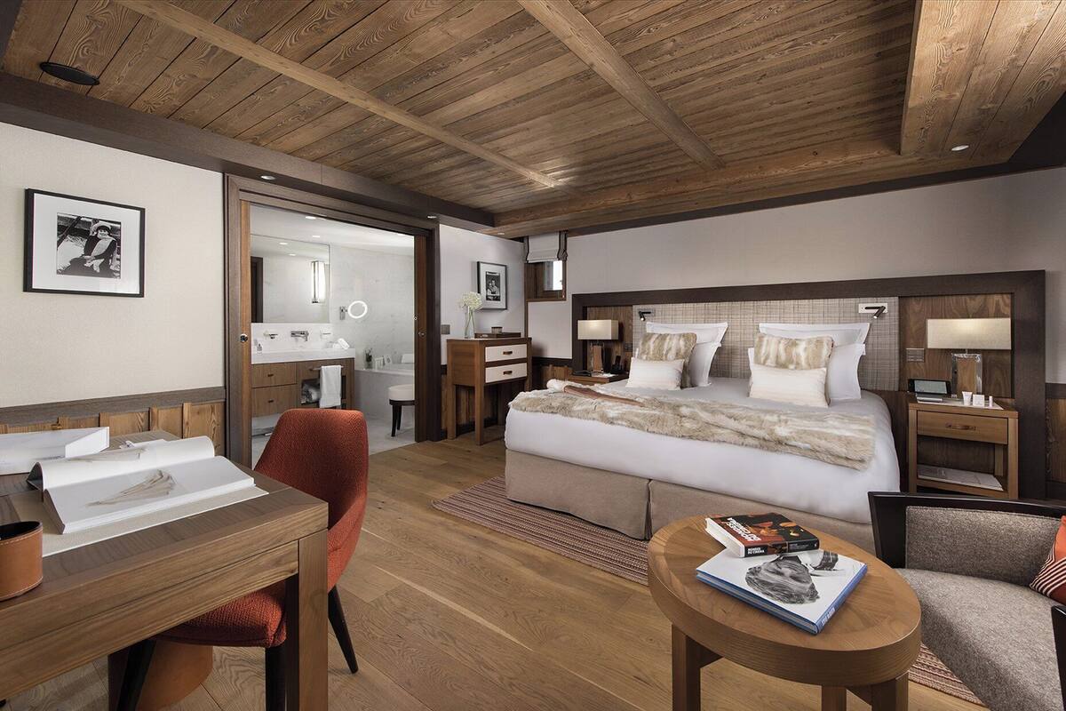 Habitación de hotel con techo de madera, una cama grande con ropa de cama blanca, un escritorio con una silla, una mesita con un libro y un sofá.
