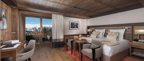 A hotel room with a wooden ceiling, a large bed with multiple pillows, a desk with a chair, a small table with a vase, and a view of snow-capped mountains through a sliding door.